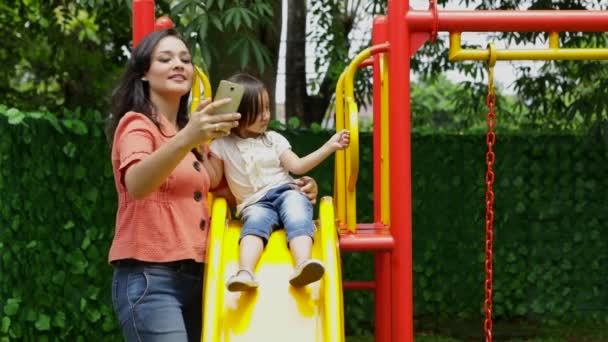 年轻的母亲和她的女儿用手机在公园的滑梯上拍自拍照片 — 图库视频影像