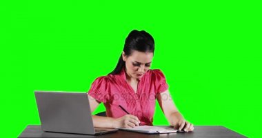 Genç işkadını iken bir kağıt yazma ve yeşil ekran arka planı 4 k çözünürlükte çekilen bir dizüstü bilgisayar bakarak masada çalışma