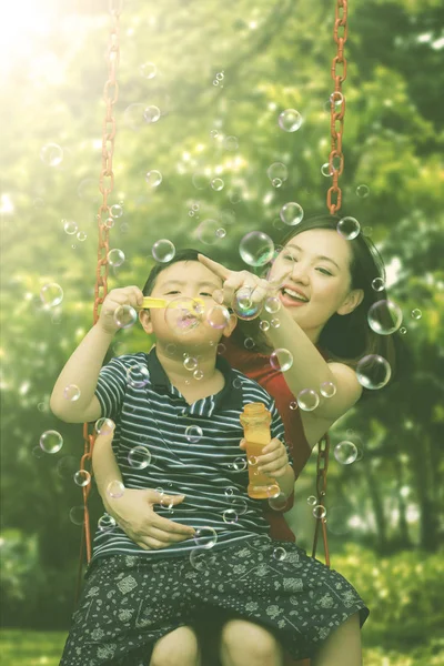 Criança com mãe jogando bolhas de sabão no parque — Fotografia de Stock