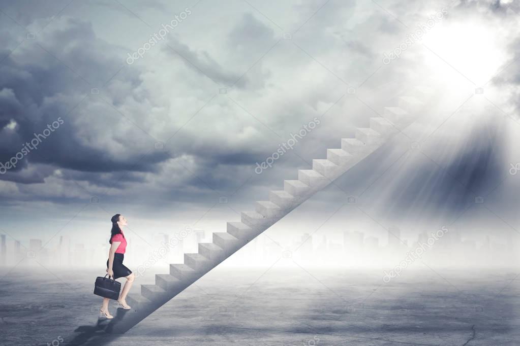 Businesswoman walking up staircase to door in sky with bright light shining down