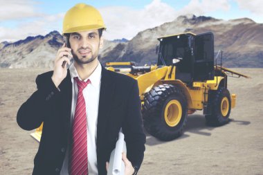 Erkek mimar önünde bir Ekskavatör inşaat sitesinde bir telefon görüşmesi yaparken laptop ile çalışma