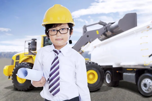 Carino ragazzo con casco di sicurezza e stampa blu di fronte a un camion di carico escavatore con terreno in un cantiere — Foto Stock