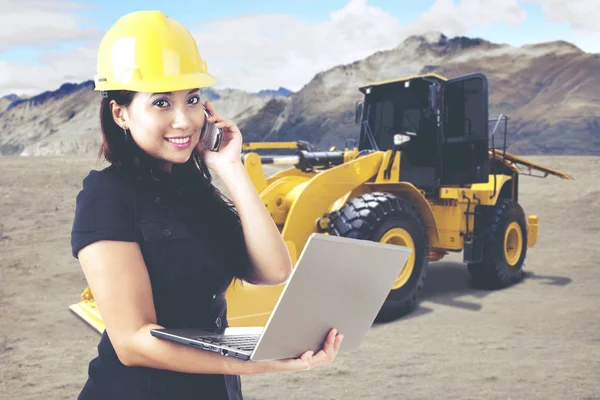 Młody architekt kobiece pracę z laptopa podczas rozmowy telefonicznej przed koparki na budowie — Zdjęcie stockowe