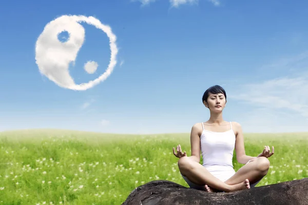 Mulher asiática meditando ao ar livre na rocha no prado com yin yang nuvens símbolo no céu — Fotografia de Stock