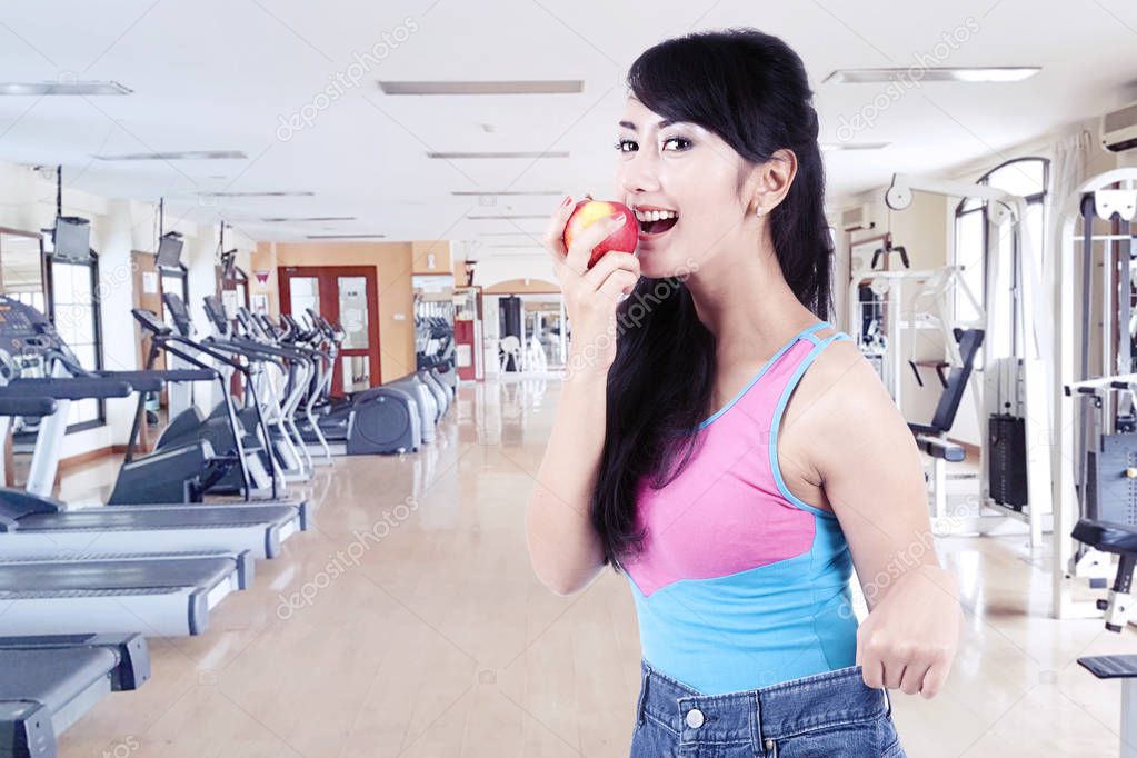 Slim young Asian woman in big jeans showing successful weight loss while eating apple