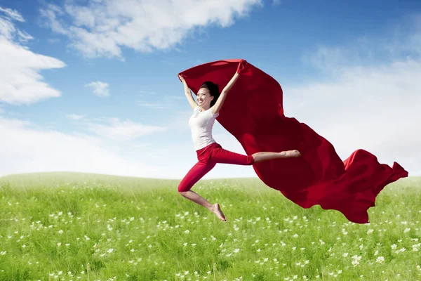 Asian woman ballerina holding red fabric making a big jump on blossom meadow. — Stock Photo, Image