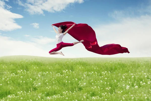 Asian woman ballerina holding red fabric making a big jump on blossom meadow. — Stock Photo, Image