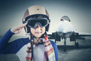 Portre sevimli küçük erkek pilot bir jet uçağı arka plan ile bir saygılı el hareketi vererek