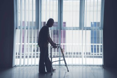 Pencerenin yakınındaki bir walker ile yaşlı adam girer