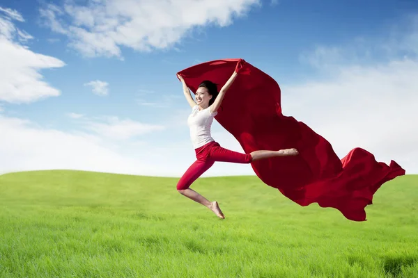 Asian woman ballerina holding red fabric making a big jump on green meadow. — Stock Photo, Image