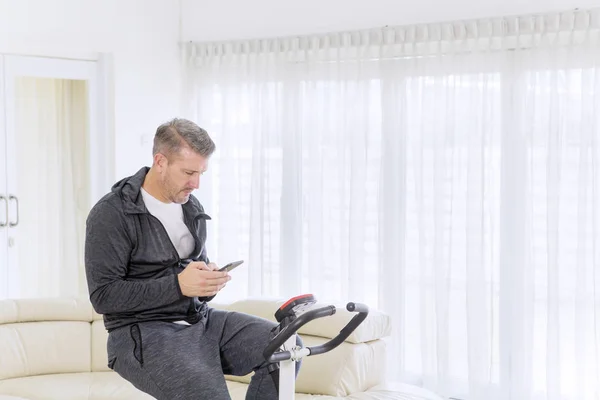 Mature man using a phone on a spin bike at home — Stock Photo, Image
