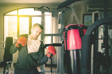 Beyaz adam spor salonunda bir boks çantasına asılıyor.