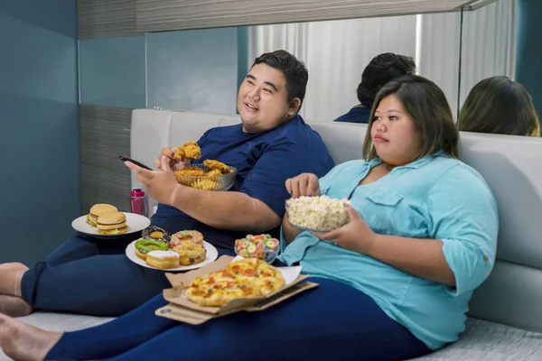 Pareja gorda sosteniendo comida chatarra mientras ve la televisión — Foto de Stock