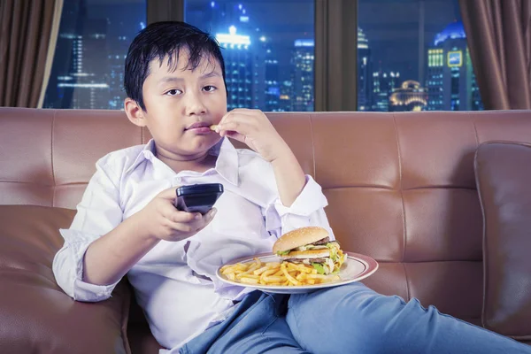 Niño comiendo hamburguesa y papas fritas en el sofá — Foto de Stock