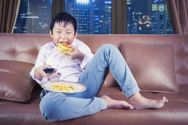 Głodny chłopiec korzystających burger siedząc na kanapie — Zdjęcie stockowe