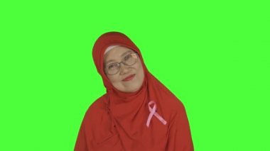 Stüdyoda dikilirken kameraya gülümseyen pembe kurdeleli yaşlı Müslüman kadın. Yeşil ekran arkaplanlı 4k çözünürlükte çekilen