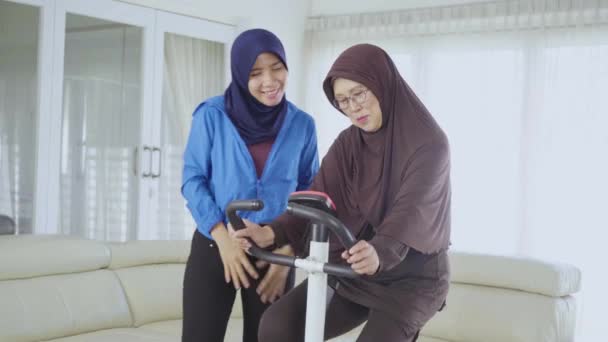 古いイスラム教徒の女性は自宅のリビングルームで娘と一緒に運動自転車でトレーニングをしています 4Kの解像度で撮影 — ストック動画