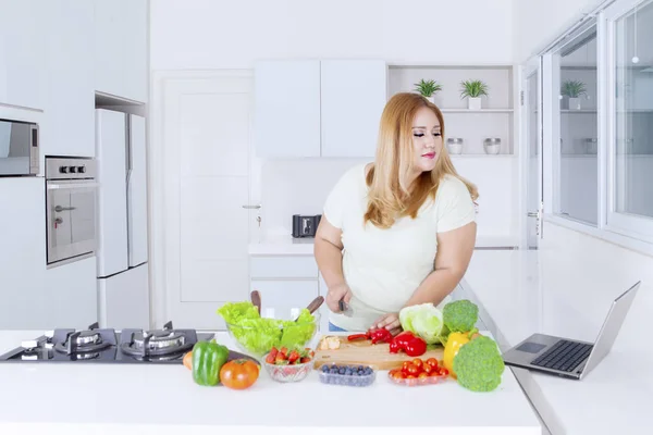 Obese woman making salad while looking at laptop