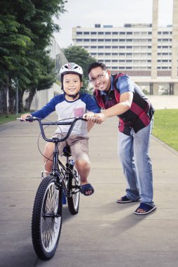 Yakışıklı Asyalı adam parkta bisiklet sürerken kameraya gülümsüyor.