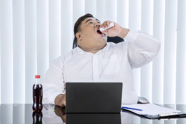 一位肥胖的亚洲商人一边在办公室的笔记本电脑上工作 一边吞吞吐吐着巧克力糖果瓶的画像 — 图库照片