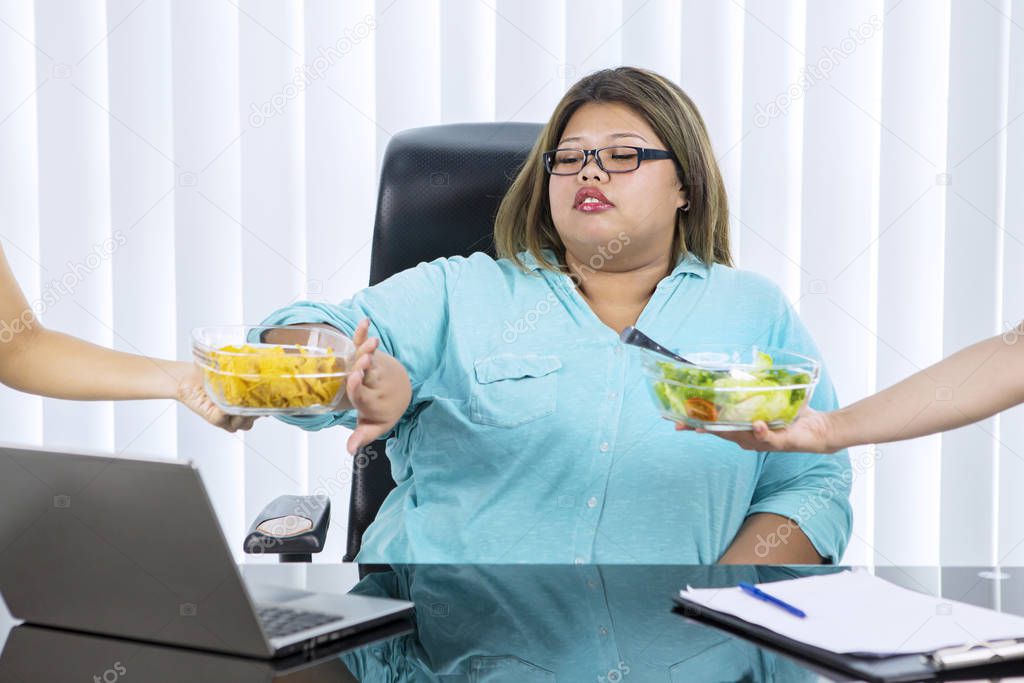 Portrait of fat Asian woman wearing formal attire, while preferring lettuce salad over chips in her office