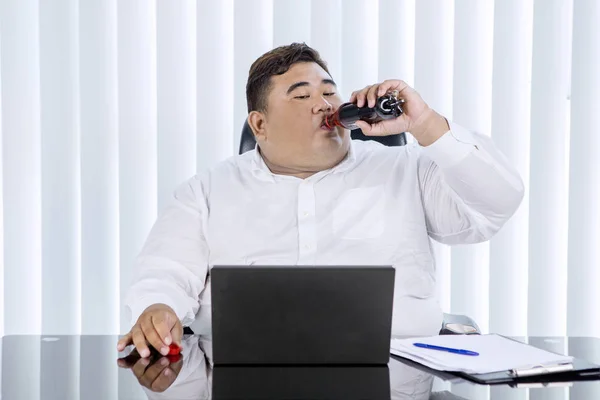 一位肥胖的亚洲商人一边在办公室里的笔记本电脑上工作 一边吞下可乐瓶的画像 — 图库照片