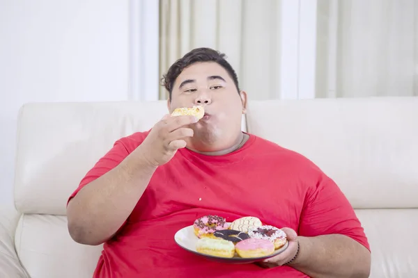 彼のリビングルームでドーナツを食べながら カメラを見て脂肪アジアの男の肖像画 — ストック写真