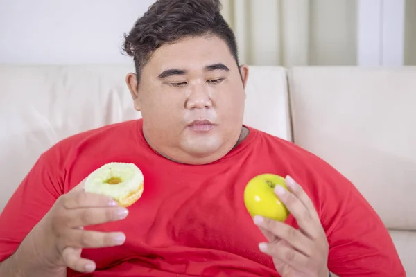 リビングルームでアップル ドーナツを選択することと混同しながらソファに座って脂肪アジアの男の肖像画 — ストック写真