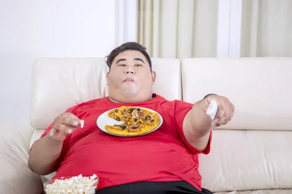 胸にピザのプレートを持ついくつかのポップコーンをつかむ太ったアジアの男の肖像 彼のリビングルームで彼のテレビのリモコンを押しながら — ストック写真