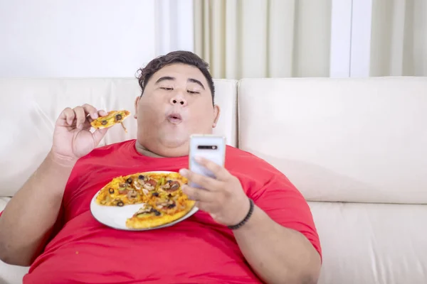 自宅のリビングルームでピザを噛みながら 彼のスマートフォンを使用して脂肪アジアのママの肖像画 — ストック写真
