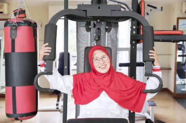 Spor salonunda egzersiz yaparken kameraya gülümseyen sağlıklı Müslüman bir kadının portresi.
