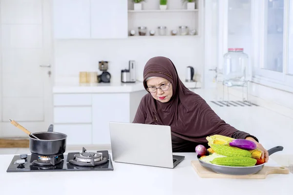 高级穆斯林妇女坐在厨房里看笔记本电脑食谱的肖像 — 图库照片