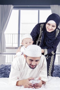 Genç Müslüman baba evdeki yatakta oğluyla oynarken cep telefonu kullanıyor.