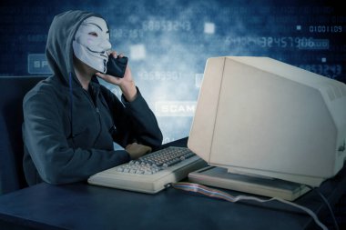 JAKARTA, Indonesia - March 10, 2020: Hacker talking on mobile phone with vendetta mask and old computer on table