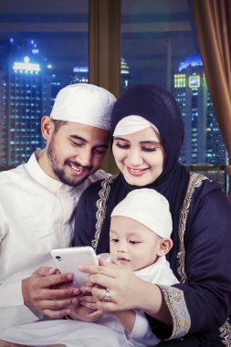 Genç Müslüman ebeveynler ve oğulları gece pencerenin yanında otururken cep telefonu kullanıyorlar.