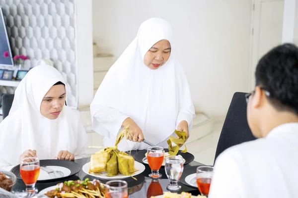 高齢者のアジアのイスラム教徒の女性の肖像彼女の家族が食べるためにケツアーを切断しながら 彼らのダイニングルームでIdul Fitriを祝います — ストック写真
