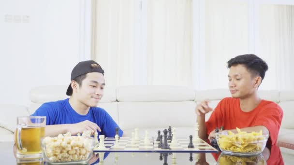 テーブルの上で食べ物や飲み物とリビングルームに座っている間 一緒にチェスをしている2人の若い男性の肖像画 — ストック動画