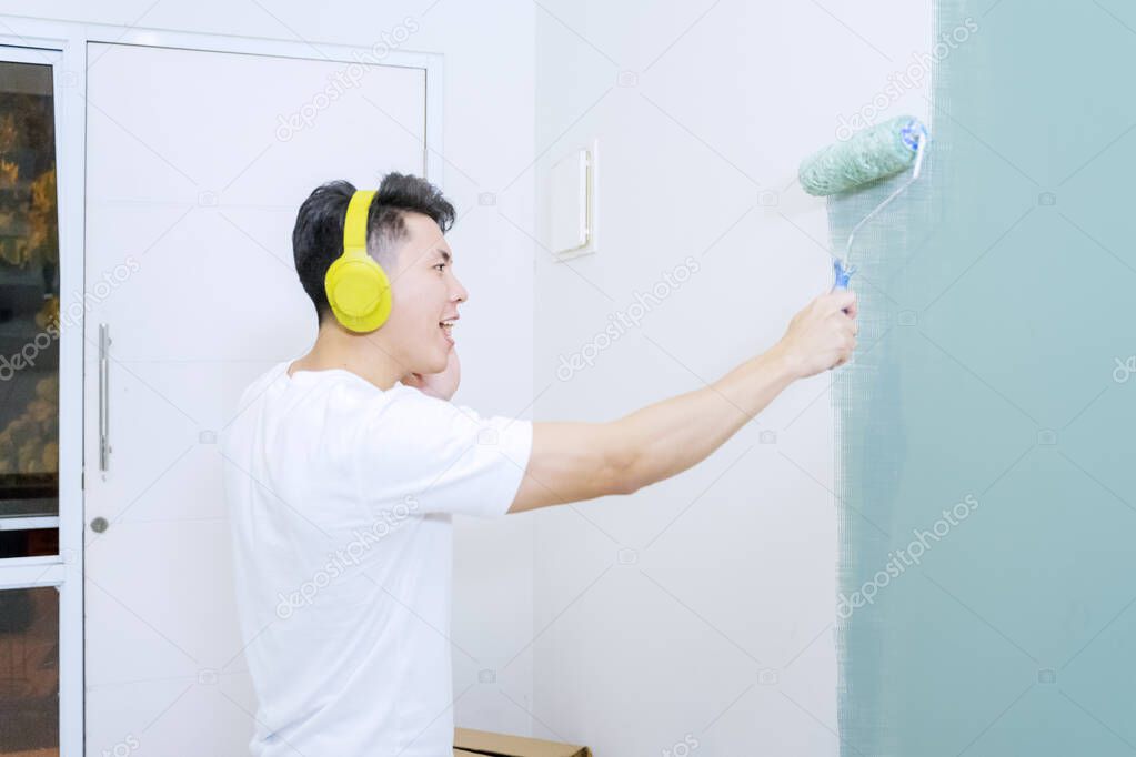 Happy young man painting interior wall with paint roller while enjoying music in new house