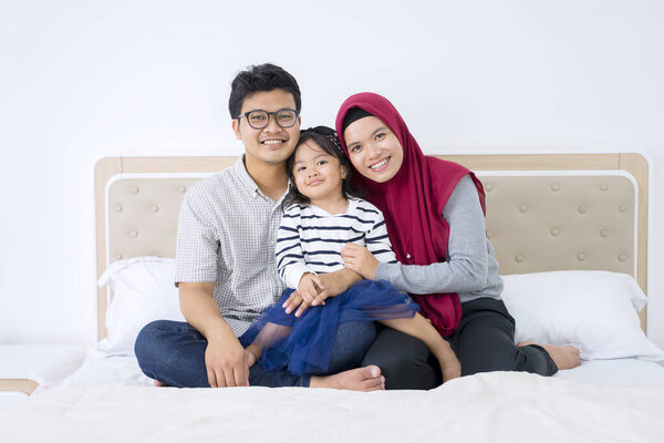 Portrait of young muslim parents and cute child smiling at camera while sitting on the bed together 