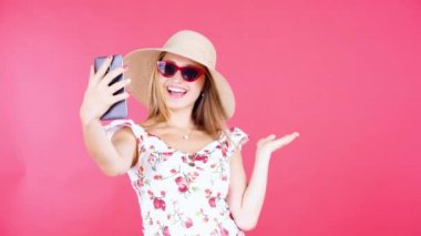 Stüdyoda kırmızı arka planda güneş gözlüğü ve şapka takıp selfie çeken güzel bir genç kadın.