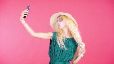Stüdyoda kırmızı arka planda güneş gözlüğü ve şapka takıp selfie çeken güzel bir genç kadın.
