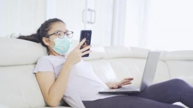Evde karantina sırasında dizüstü bilgisayar ve cep telefonu kullanan genç bir kadın.