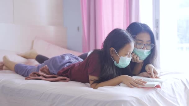 若い母親と彼女の娘の肖像は 医療マスクを身に着けている間 ベッドの上で本を読んで 家で安全の概念 — ストック動画