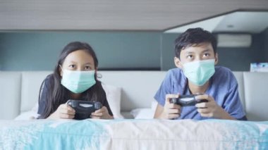 Yatakta tıbbi maske takıp oyun oynayan kız ve erkek kardeşlerin portresi.