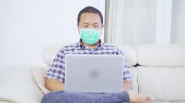 Adam maske takıyor ve dizüstü bilgisayarla koltukta çalışıyor.