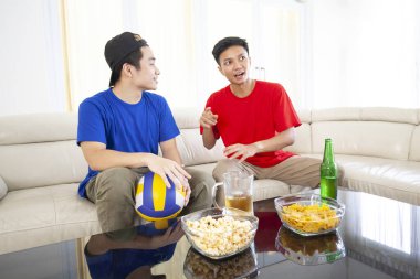 Kanepede yan yana otururken voleybol yarışmasından bahseden iki Asyalı adamın portresi. 