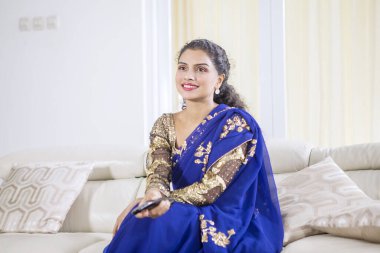 Oturma odasında mavi sari giyip televizyon izleyen çekici bir kadının portresi. 