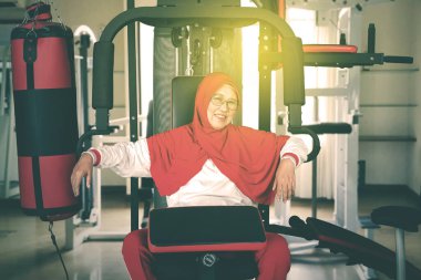 Yaşlı Müslüman kadının portresi spor aletlerinin üzerinde otururken kameraya gülümsüyor ve dinleniyor.  