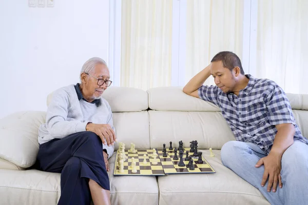 Portrait of old man and his son playing chess together in living room while thinking game strategy