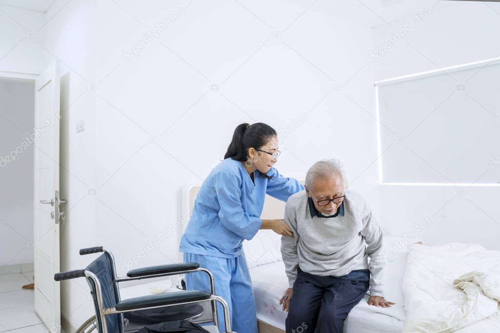 Portrait of young nurse helping senior man to sit on the bed while standing in the bedroom